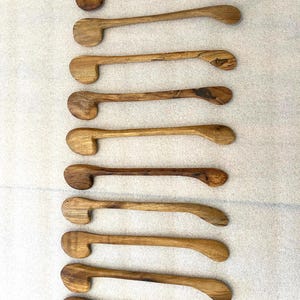 May include: A collection of nine wooden spoons, each with a unique grain pattern and a rounded bowl. The spoons have long, slender handles and are arranged vertically against a light background. The wood tones vary from light to dark brown.