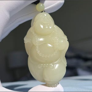 May include: A light green jade pendant in the shape of a smiling Buddha. The pendant features a beaded necklace and a small ball in one hand. The Buddha is suspended from a black cord with a small jade bead.
