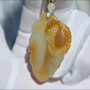 May include: A pendant featuring a leaf and fruit design, crafted from translucent yellow and orange material. The pendant is suspended from a silver-colored cord and a small bead. The pendant is held by a gloved hand.