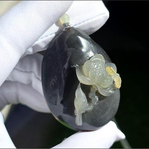 May include: A teardrop-shaped dark charcoal pendant featuring a carved light green floral motif with a small yellow accent. The smooth, polished stone has a small translucent bead at the top, creating a contrast between the dark base and the light carving.