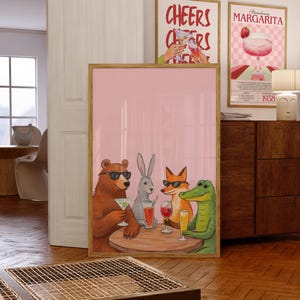 May include: A framed print featuring a bear, rabbit, fox, and alligator wearing sunglasses and enjoying drinks at a table. The print has a pink background and is in a wooden frame. Two other framed prints are visible, one with the text "Cheers" and the other with "Strawberry Margarita".