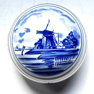 May include: Round, white ceramic trinket box with a blue and white windmill design on the lid. The lid features a detailed illustration of a Dutch windmill, water, and a small house. The box has a simple, elegant design.