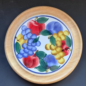 May include: Round wooden serving tray with a hand-painted ceramic center. The ceramic features a colorful design of red apples, blue grapes, yellow pears, and purple plums with green leaves. The tray has a light brown wooden rim.