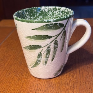 May include: Cream-colored ceramic mug with a white handle and a green speckled rim. The mug features a hand-painted olive branch design in shades of green. The mug is sitting on a wooden surface.
