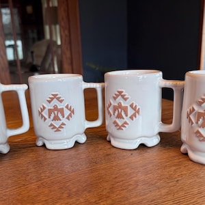 May include: Four white ceramic mugs with a glossy finish, each featuring a raised, light brown geometric pattern centered on the front. The design depicts a stylized bird motif surrounded by diamond-shaped accents. Each mug has a sturdy handle.
