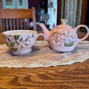 May include: A pink ceramic teacup and teapot set. The set features a floral design with white and purple flowers and green leaves. The teacup has a handle and sits on a small base. The teapot has a lid and a curved spout.