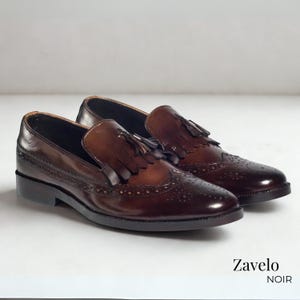 Op de afbeelding: Een paar bruine leren loafers met decoratieve perforaties en kwastjes. De schoenen hebben een glanzende afwerking en een klassiek ontwerp, perfect voor formele of zakelijke kleding. De merknaam "Zavelo NOIR" is zichtbaar.