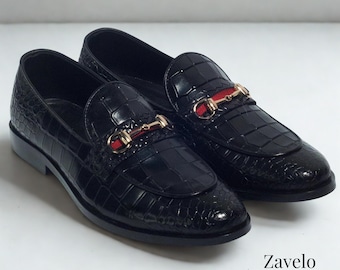 May include: A pair of black leather loafers with a crocodile texture. Each shoe features a gold-colored metal accent with a red stripe. The brand name "Zavelo Noir" is printed in the bottom right corner.