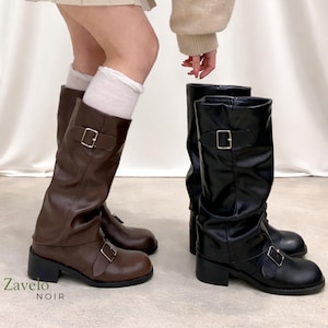 May include: Two pairs of knee-high boots. One pair is brown, the other is black. Both have a buckle detail and a block heel. The brown boots are on the left, and the black boots are on the right. The text "Zavelo NOIR" is at the bottom left.