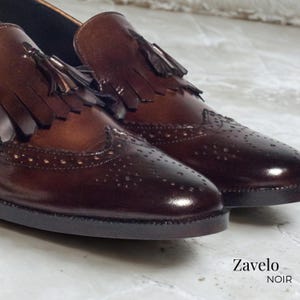 Op de afbeelding: Close-up van een paar bruine leren herenschoenen met decoratieve perforaties en kwastjes. De schoenen hebben een gepolijste afwerking en een donkere zool. De tekst "Zavelo NOIR" is zichtbaar.