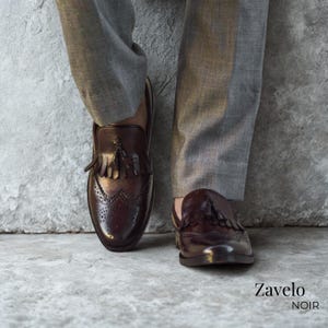 Op de afbeelding: Close-up van bruine leren loafers met kwastjes en brogue details. De schoenen worden gecombineerd met een grijze broek. De merknaam "Zavelo Noir" is rechtsonder zichtbaar.
