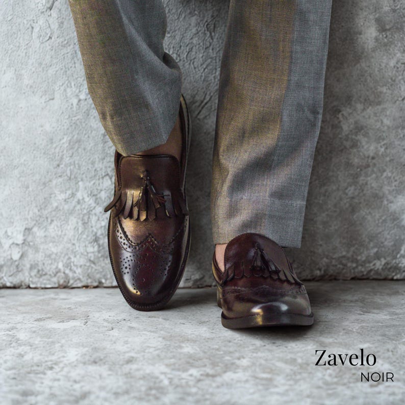 Op de afbeelding: Close-up van bruine leren loafers met kwastjes en brogue details. De schoenen worden gecombineerd met een grijze broek. De merknaam "Zavelo Noir" is rechtsonder zichtbaar.