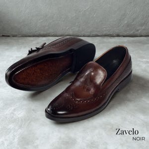 Op de afbeelding: Een paar donkerbruine leren loafers met decoratieve perforaties en een glanzende afwerking. De schoenen hebben een klassiek ontwerp met een ronde neus en een lage hak. De merknaam "Zavelo NOIR" is zichtbaar.