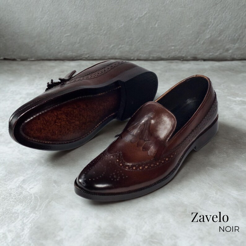 Op de afbeelding: Een paar donkerbruine leren loafers met decoratieve perforaties en een glanzende afwerking. De schoenen hebben een klassiek ontwerp met een ronde neus en een lage hak. De merknaam "Zavelo NOIR" is zichtbaar.