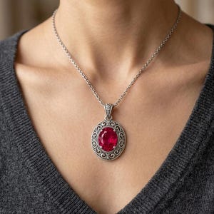 May include: A silver pendant necklace featuring a large, oval, ruby-red gemstone. The pendant is set in an ornate, circular frame with a detailed, swirling design. The necklace is worn on a person.