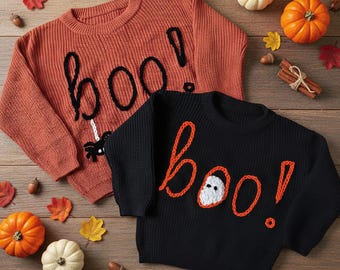 May include: Two Halloween-themed sweaters. One is burnt orange with a black "boo!" design and a spider. The other is black with an orange "boo!" design and a ghost. Pumpkins, leaves, and acorns are scattered around.