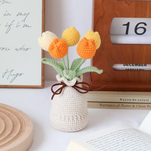May include: A crocheted bouquet of tulips in a cream-colored vase. The flowers are in shades of yellow, orange, and cream, with green leaves. A brown ribbon ties the vase. A wooden calendar shows Saturday, the 16th.