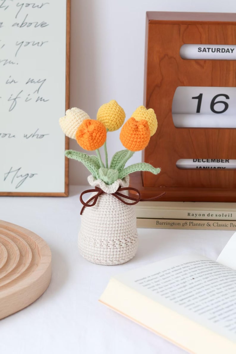 May include: A crocheted bouquet of tulips in a cream-colored vase. The flowers are in shades of yellow, orange, and cream, with green leaves. A brown ribbon ties the vase. A wooden calendar shows Saturday, the 16th.