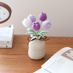 May include: A crocheted bouquet of tulips in a small, beige vase. The flowers are in shades of white, lavender, and purple, with green leaves. The vase is tied with a brown ribbon. The arrangement sits on a wooden surface.