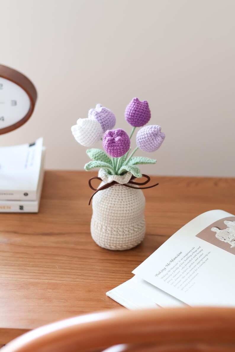 May include: A crocheted bouquet of tulips in a small, beige vase. The flowers are in shades of white, lavender, and purple, with green leaves. The vase is tied with a brown ribbon. The arrangement sits on a wooden surface.