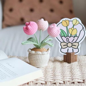 May include: A crocheted flower arrangement featuring three pink tulips in a small, off-white pot. The arrangement sits on a woven surface next to an open book and a floral illustration on a wooden stand. The background includes a soft, neutral-toned pillow.