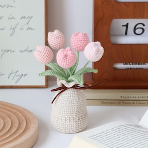 May include: A crocheted bouquet of pink tulips in a cream-colored vase tied with a brown ribbon. The handmade flowers have green stems and leaves. A wooden tray, a book, and a calendar showing Saturday, December 16 are in the background.