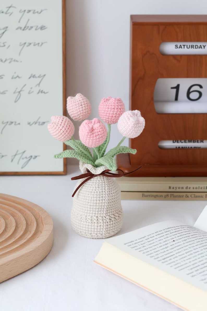 May include: A crocheted bouquet of pink tulips in a cream-colored vase tied with a brown ribbon. The handmade flowers have green stems and leaves. A wooden tray, a book, and a calendar showing Saturday, December 16 are in the background.