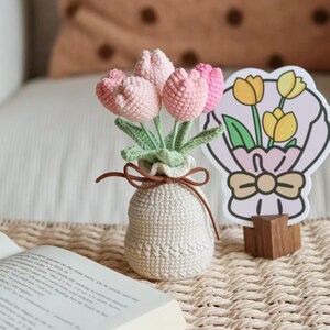 May include: A crocheted bouquet of pink tulips in a small, off-white vase tied with a brown ribbon. A decorative card with a tulip bouquet illustration stands nearby. The arrangement sits on a woven mat.