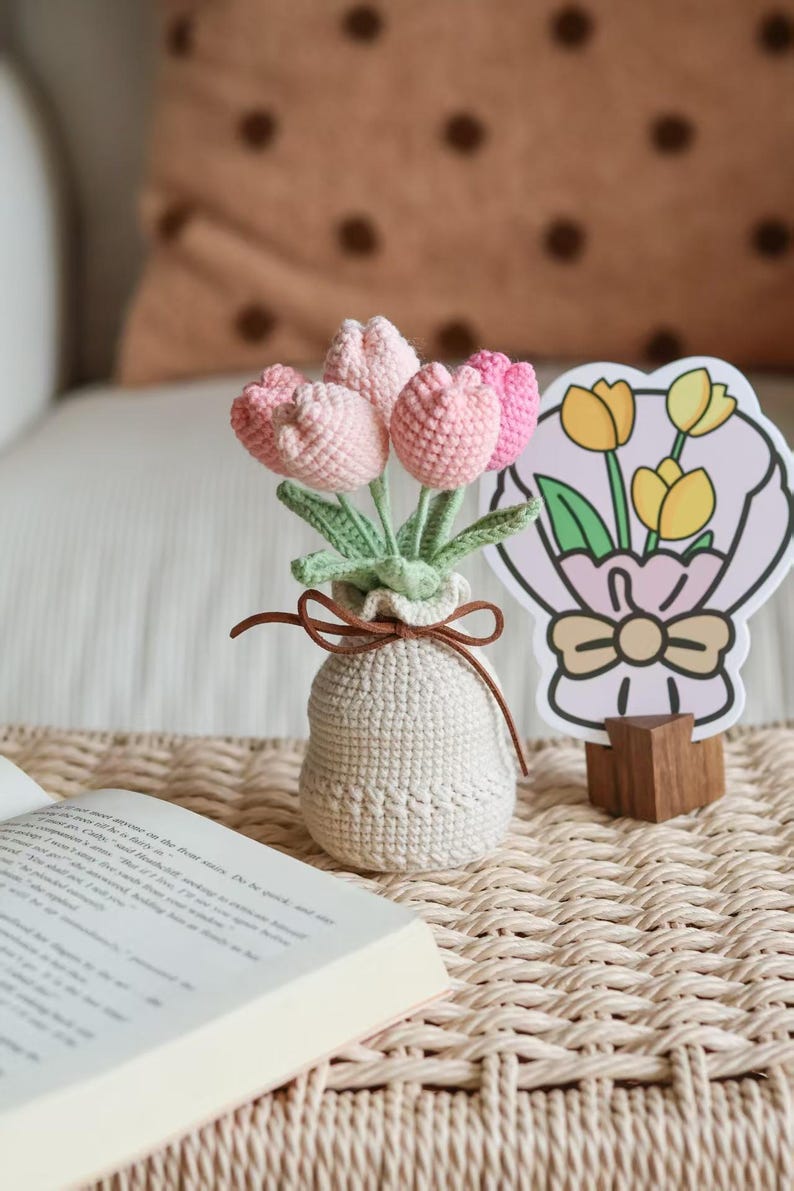 May include: A crocheted bouquet of pink tulips in a small, off-white vase tied with a brown ribbon. A decorative card with a tulip bouquet illustration stands nearby. The arrangement sits on a woven mat.