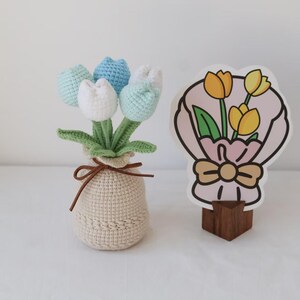May include: A crocheted tulip bouquet in a beige pot with a brown tie. The tulips are in shades of white, blue, and green. A decorative card with a bouquet of yellow tulips is on a wooden stand.