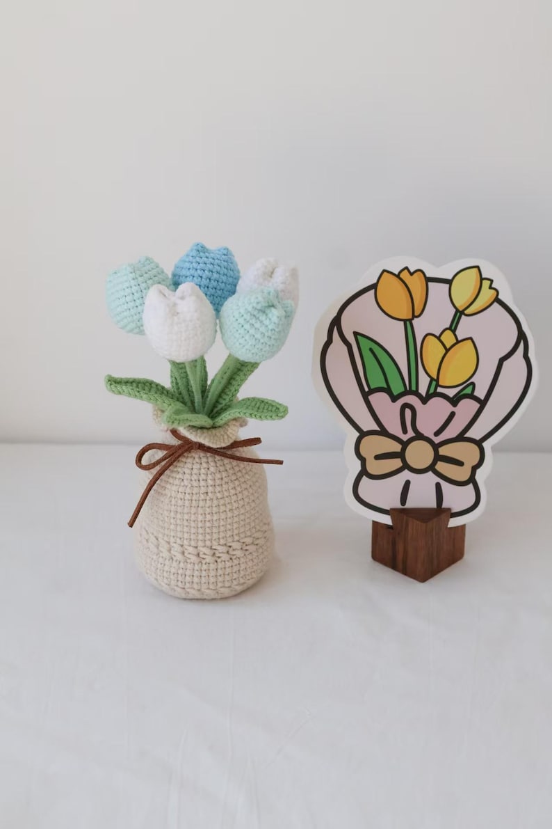 May include: A crocheted tulip bouquet in a beige pot with a brown tie. The tulips are in shades of white, blue, and green. A decorative card with a bouquet of yellow tulips is on a wooden stand.