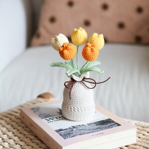 May include: A crocheted bouquet of tulips in a small white vase, tied with a brown ribbon. The tulips are in shades of yellow, orange, and cream, with green leaves. The vase sits on a book with a pink cover.