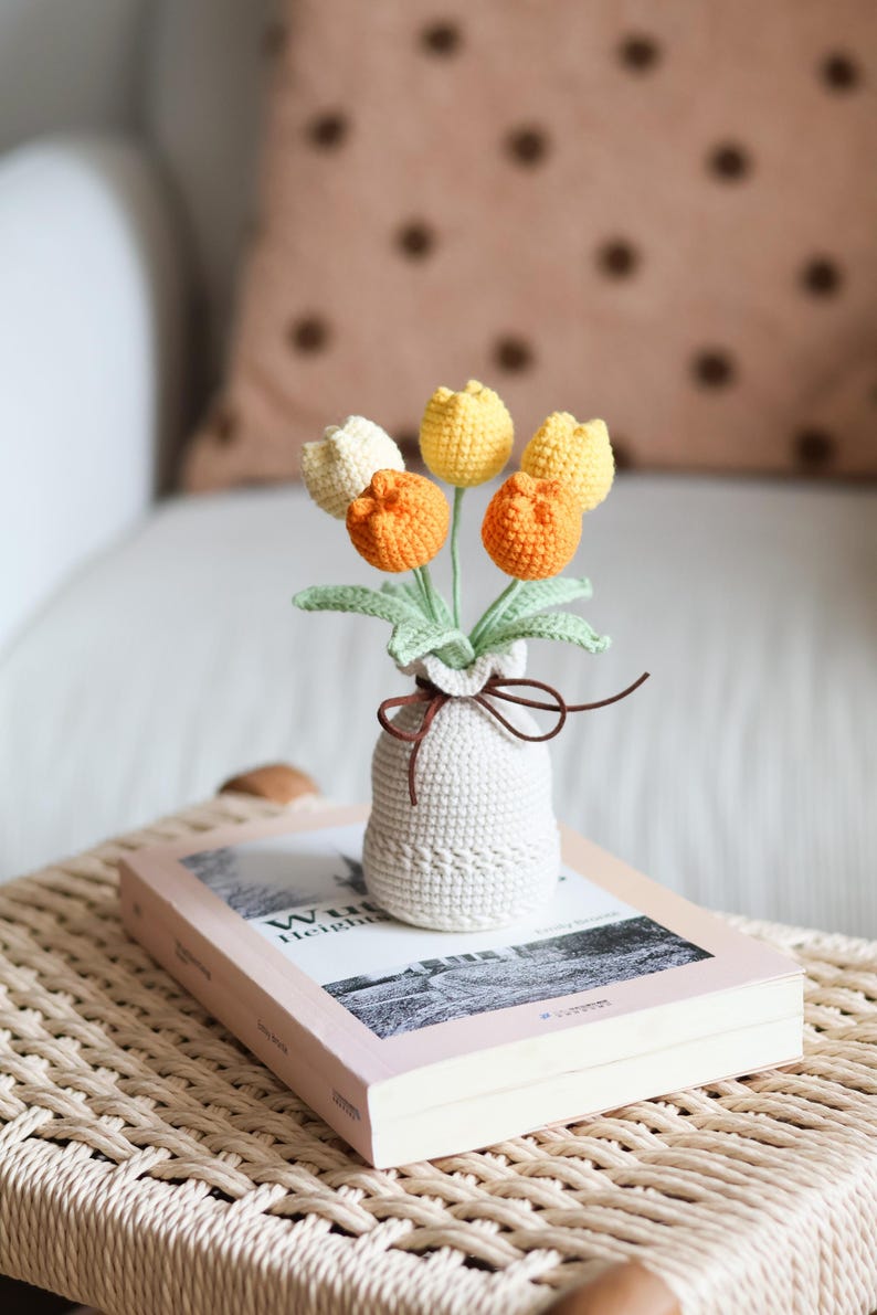 May include: A crocheted bouquet of tulips in a small white vase, tied with a brown ribbon. The tulips are in shades of yellow, orange, and cream, with green leaves. The vase sits on a book with a pink cover.