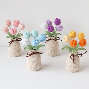 May include: A collection of handmade crochet tulip bouquets in various colors, including pink, blue, purple, and yellow. Each bouquet is arranged in a beige crocheted vase and tied with a brown ribbon. The flowers have green leaves.