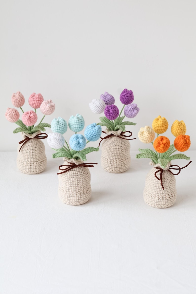 May include: A collection of handmade crochet tulip bouquets in various colors, including pink, blue, purple, and yellow. Each bouquet is arranged in a beige crocheted vase and tied with a brown ribbon. The flowers have green leaves.