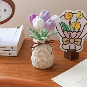 May include: A crocheted bouquet of tulips in a beige vase tied with a brown ribbon. The flowers are white and purple, with green stems and leaves. A decorative tulip-themed card is in the background.