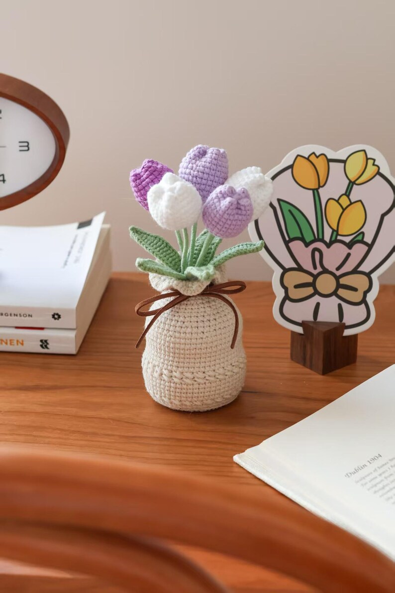 May include: A crocheted bouquet of tulips in a beige vase tied with a brown ribbon. The flowers are white and purple, with green stems and leaves. A decorative tulip-themed card is in the background.