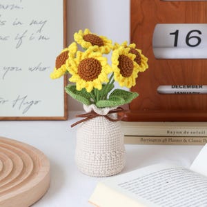 May include: A crocheted sunflower bouquet in a beige vase. The sunflowers have yellow petals and brown centers, with green leaves. The vase is tied with a brown string. A wooden calendar shows Saturday, December 16.