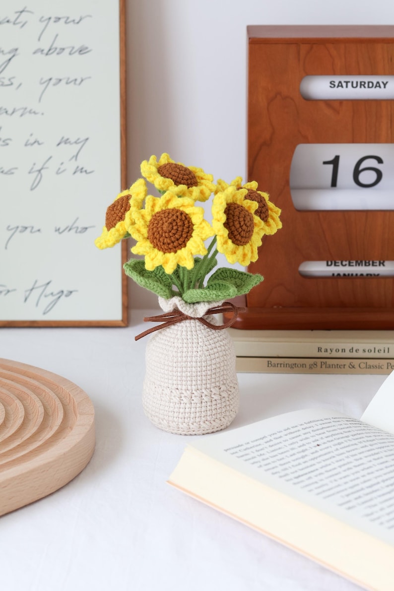 May include: A crocheted sunflower bouquet in a beige vase. The sunflowers have yellow petals and brown centers, with green leaves. The vase is tied with a brown string. A wooden calendar shows Saturday, December 16.