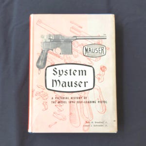 Puede incluir: Un libro antiguo titulado "System Mauser" con una historia ilustrada de la pistola semiautomática Modelo 1896. La portada presenta una ilustración detallada de una pistola Mauser y el logotipo "MAUSER".