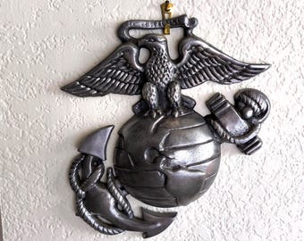 May include: A detailed, metallic emblem featuring an eagle perched atop a globe, with an anchor and rope design. The eagle has outstretched wings, and the globe is detailed with continents. The emblem is mounted on a textured wall.