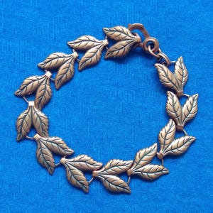 Op de afbeelding: Een bronskleurige armband met een herhalend bladerenontwerp. De bladeren zijn gedetailleerd met aderen en verbonden door kleine schakels. De armband heeft een haaksluiting. Blauwe achtergrond.
