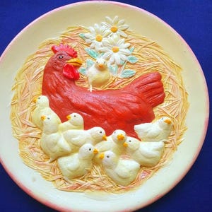 Puede incluir: Plato decorativo de cerámica con una gallina roja con pico y cresta amarillos, rodeada de pollitos amarillos y margaritas blancas. El plato tiene un fondo color crema con un borde rosa y una textura de paja.