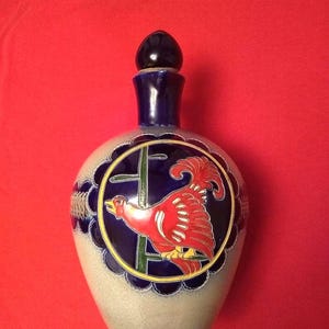 May include: A ceramic bottle with a dark blue stopper and neck. The body is light beige with a dark blue scalloped border. A red rooster is centered on the bottle within a gold and blue circle. The background is red.