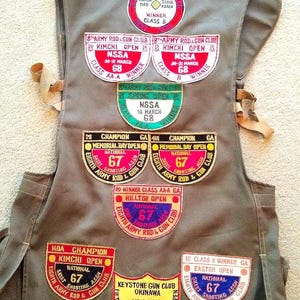 May include: A tan shooting vest with multiple patches. The patches display text such as "Winner Class B", "Kimchi Open", "Champion", and "Easter Open". The vest has adjustable straps and pockets.
