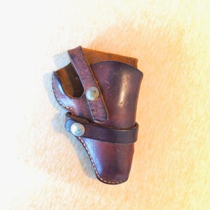 May include: A brown leather holster with a dark brown strap and silver-coloured button accents. The holster is designed to hold a firearm and features a stitched design along the edges. The leather has a worn appearance.