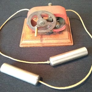 May include: An antique red and metal device with a wooden base, a large gear, and a hand crank. Two silver cylindrical objects are connected to the device by a yellow cord. The device is likely a vintage electrical or scientific instrument.
