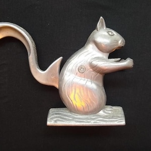May include: A silver-colored squirrel nutcracker with a curved tail and a lever. The squirrel is standing on a wooden-textured base. The nutcracker is made of metal and has a detailed design.