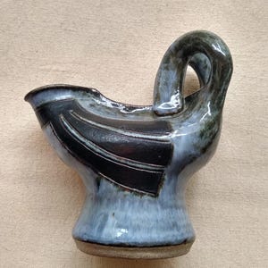 May include: Handmade ceramic creamer with a unique design. The creamer features a blue and black glaze with a curved handle and a spout. The base is a light brown color. The creamer is a functional and decorative piece for serving cream or other liquids.