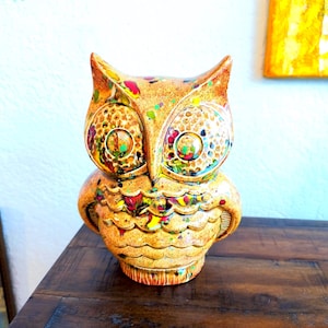 Könnte beinhalten: Eine Eulenfigur aus Keramik mit einer texturierten, goldbraunen Grundfarbe und lebendigen, mehrfarbigen Farbspritzern in Rot, Gelb, Grün und Blau. Die Eule hat große, kreisförmige Augen und detaillierte, geschwungene Federmuster.